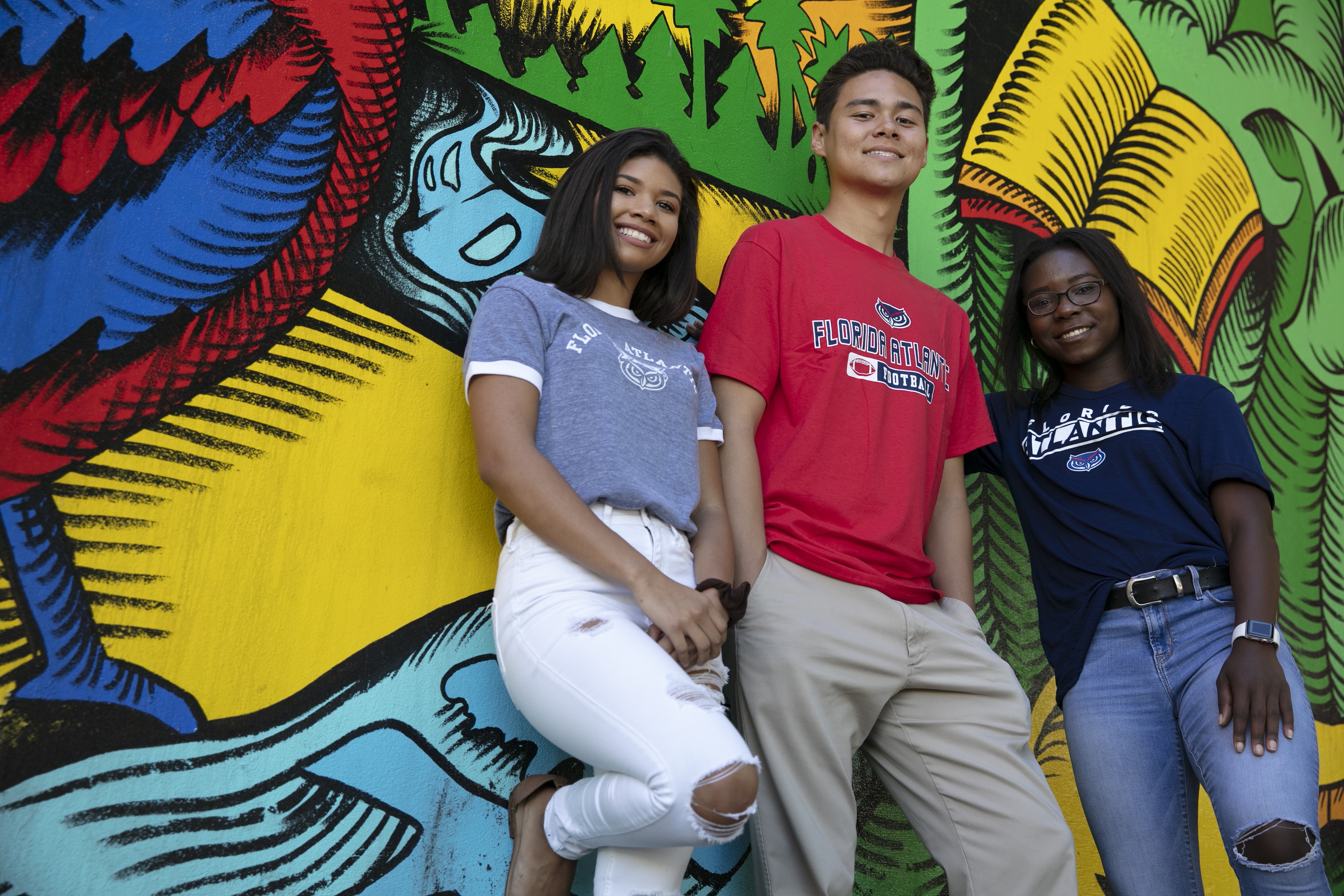 three students wearing 太阳城娱乐 shirts stand in front of a mural while smiling at the camera