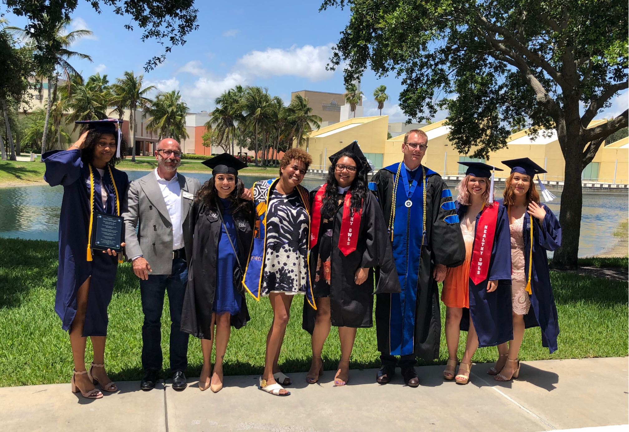 A group of eight people, many in graduation attire, standing on a walkway with a campus and palm trees in the background