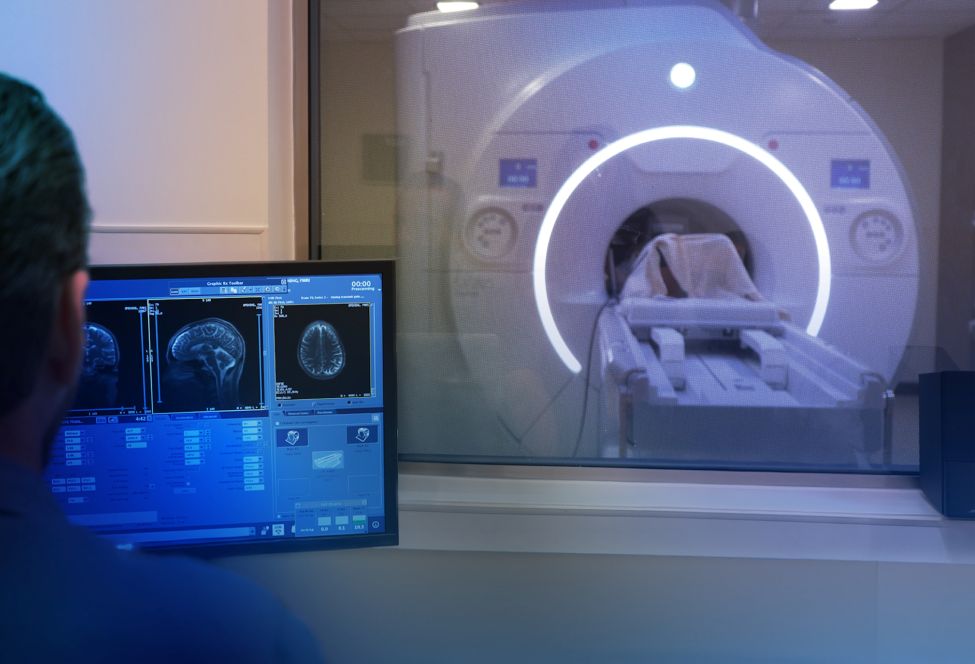Technician monitors brain MRI scans on a computer while a patient lies inside an MRI machine in a hospital imaging room.