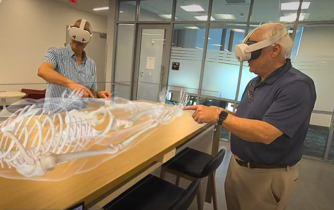 2 mean standing around a wooden table wearing white virtual reality goggles. On the table is a virtual diagram of a human skeleton.
