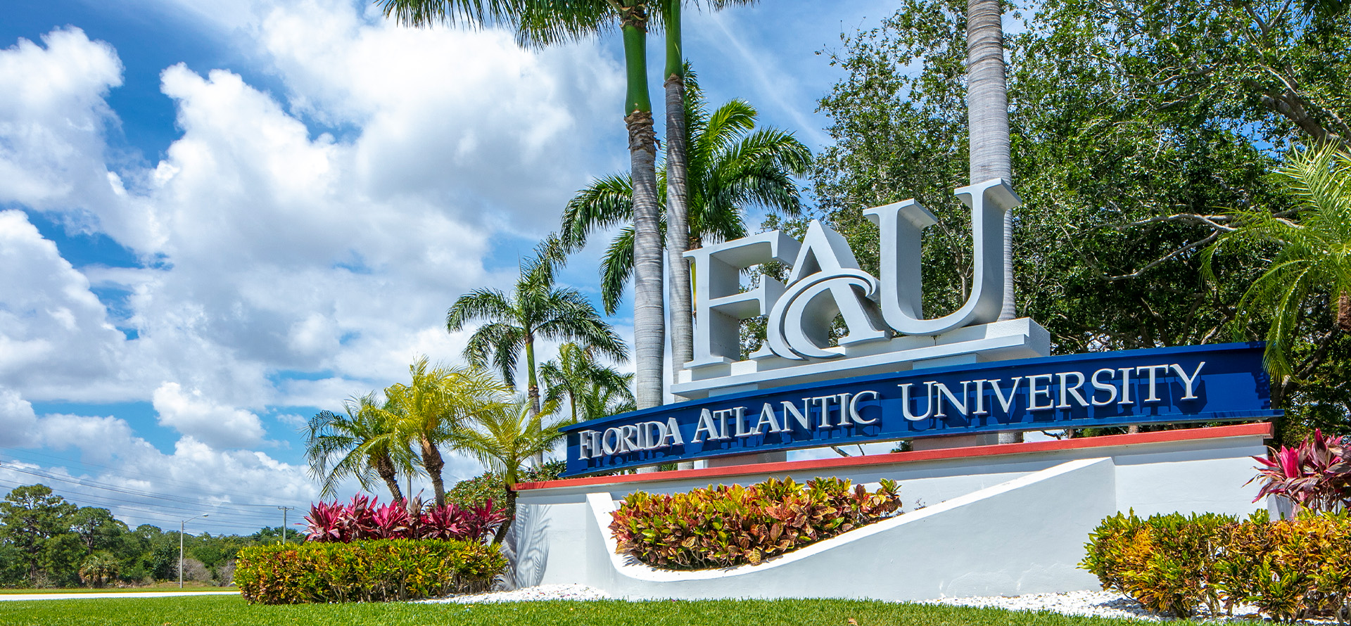 Entrance sign for Florida Atlantic University with palm trees and clouds in the background.