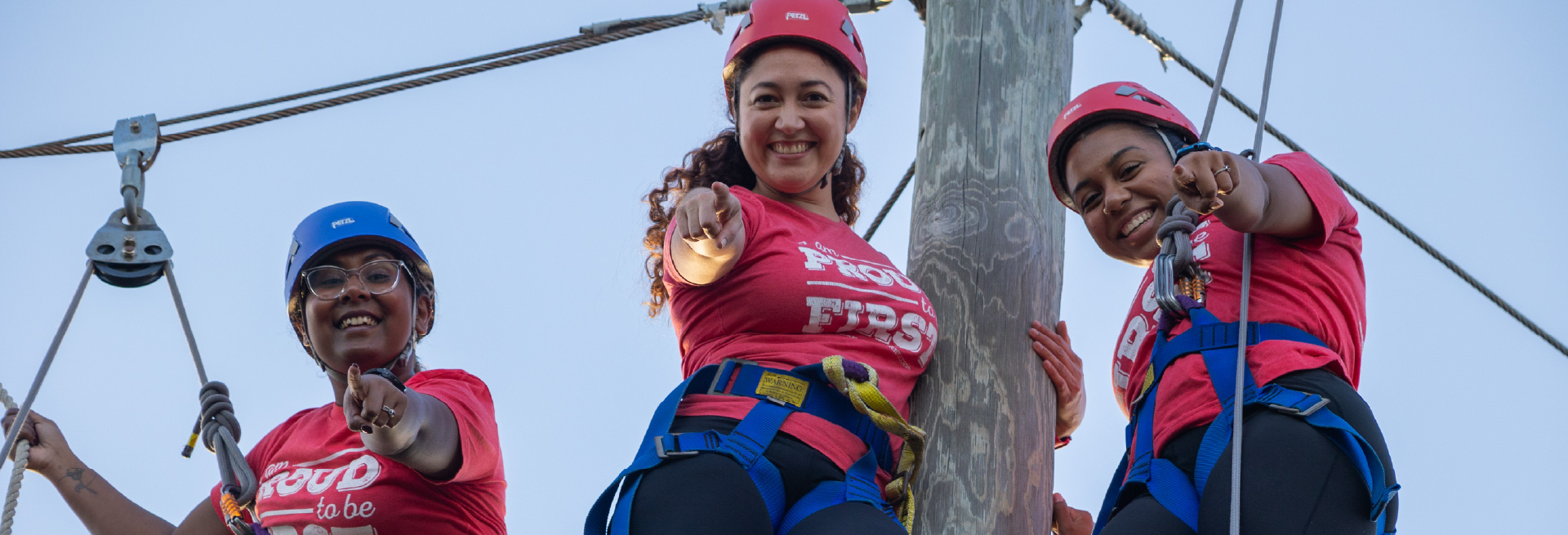 Three smiling individuals on a high ropes course, wearing helmets, harnesses, and red shirts.
