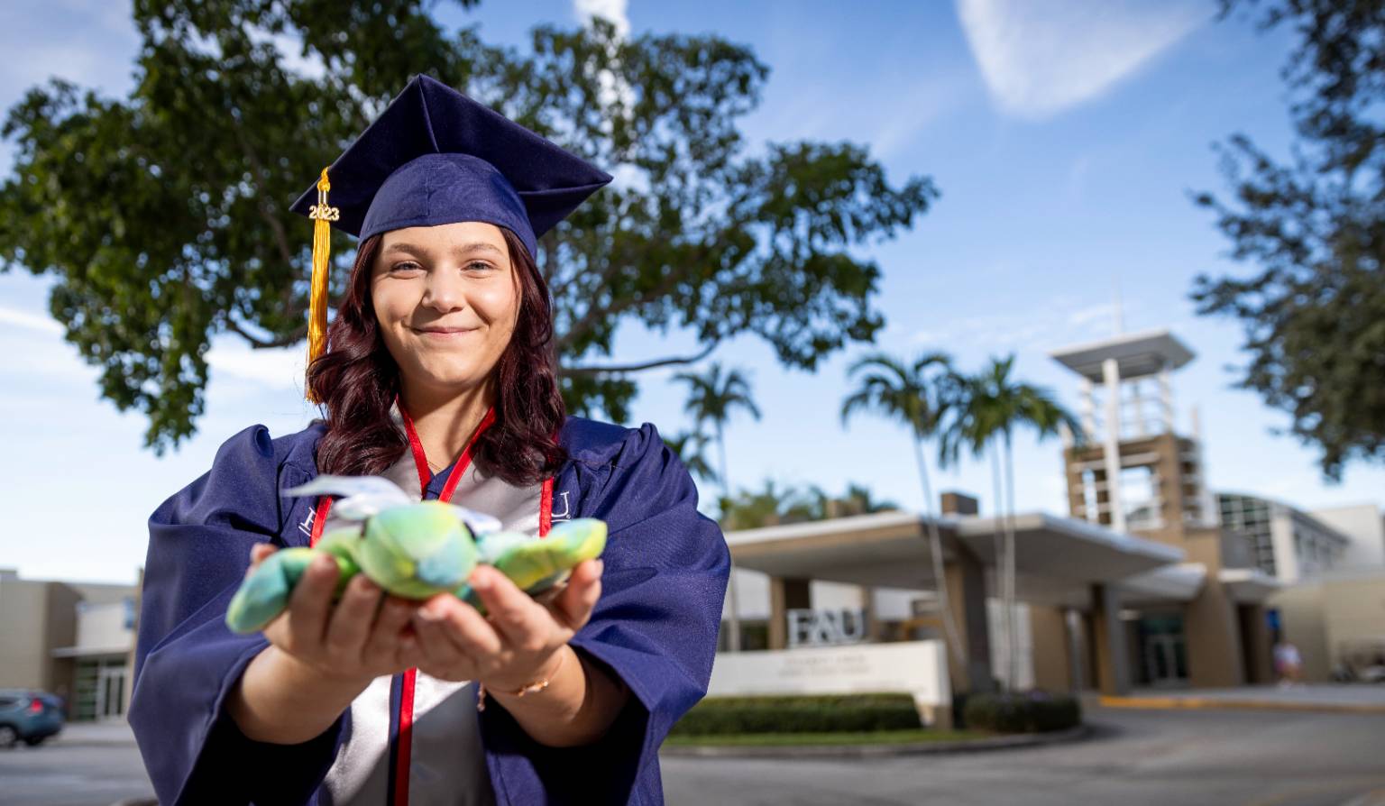 Graduate in cap and gown smiling while holding a plush sea turtle, standing outdoors at 太阳城娱乐 with palm trees and modern buildings in the background.