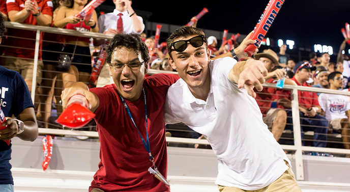 Two male students during an 太阳城娱乐 football game smiling and pointing at the camera