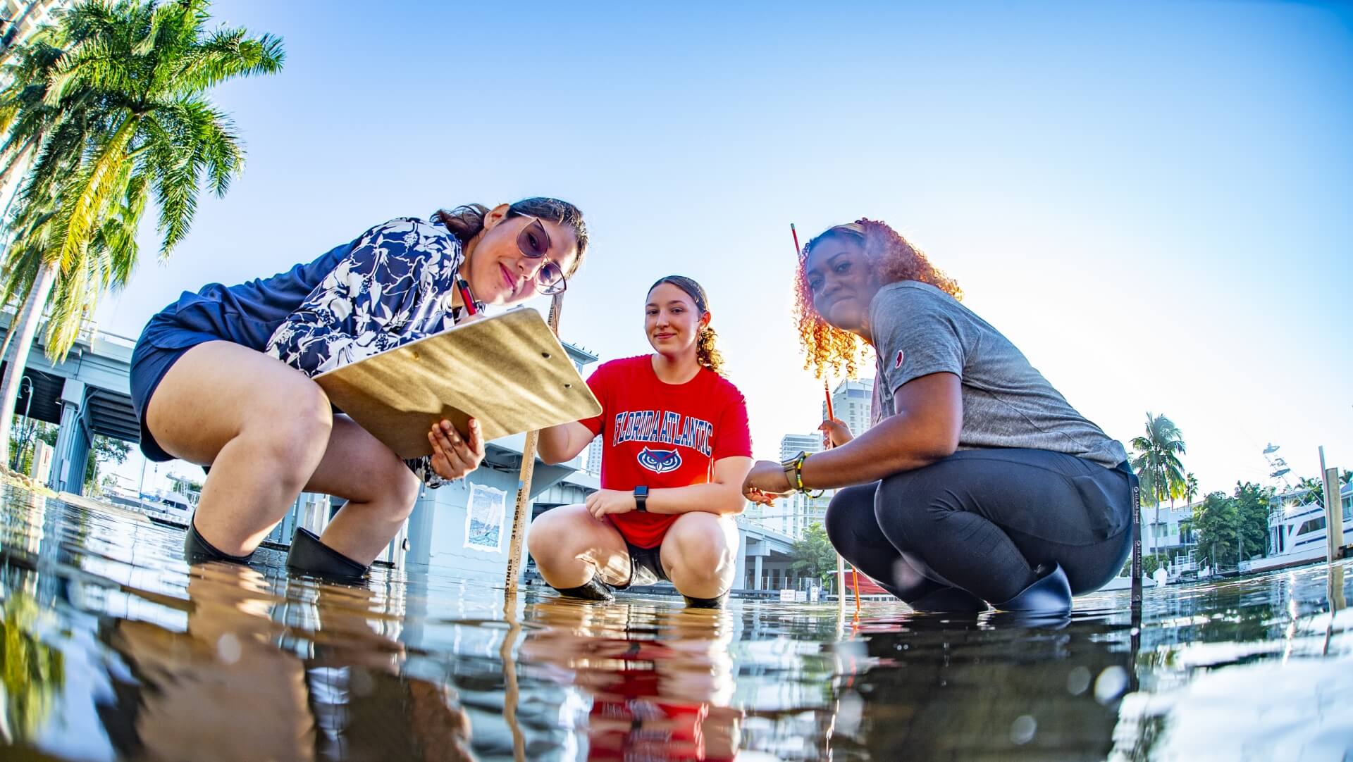 Three people conducting field 研究 in shallow water near palm trees and buildings; one holds a clipboard, another a measuring stick.