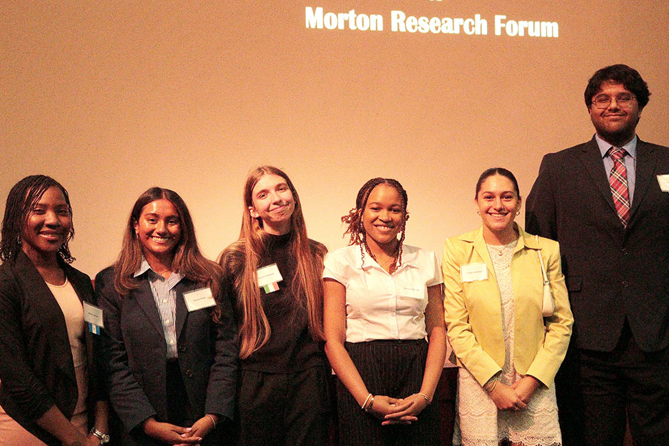 Six students posing in a line in front of a screen that reads Morton Research Forum