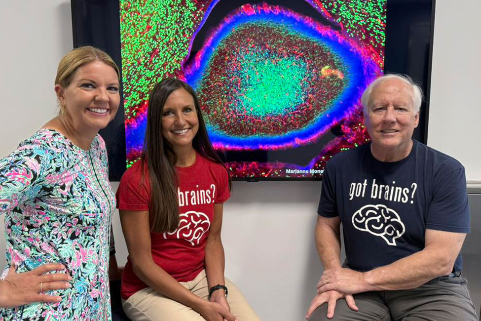 A woman stands beside another woman and man, both seated, in front of a screen with a neuron microscope image. All are smiling at the camera.