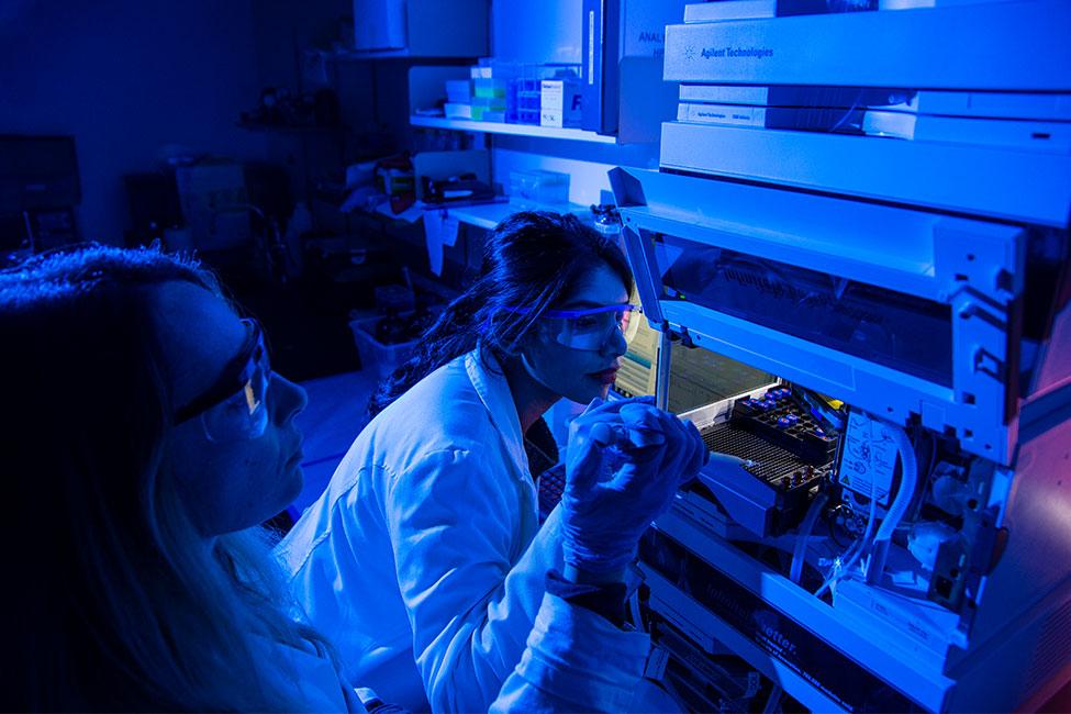 Two female scientists working with a syringe and high-tech equipment in a blue-lit research laboratory.