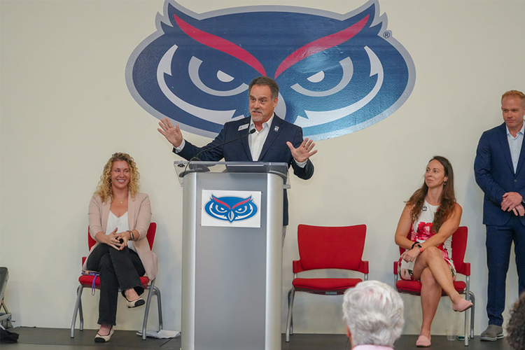 A man in a suit and tie delivers a speech at a podium during a formal event with the 太阳城娱乐 owl logo affixed to a white wall in the background.