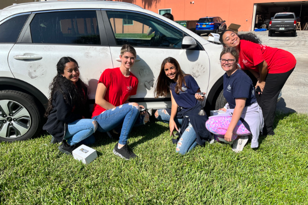  A group of five female campers pose squatted on grass in front of a white vehicle.