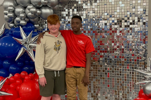 Two male campers huddle for a photo in front of a silver mirror tiled wall with red, blue and silver balloons surrounding them. 