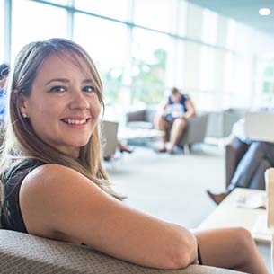 A woman smiling while seated on a couch in a bright, modern room with large windows.